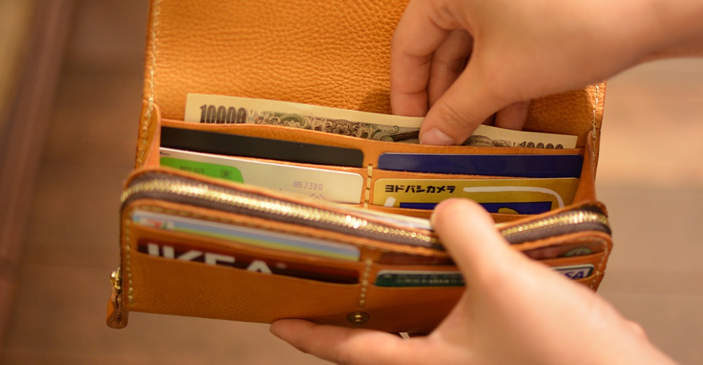 一只手正在從一個寬口的棕褐色皮革長夾中取出日圓鈔票，夾內可見多張信用卡及證件。