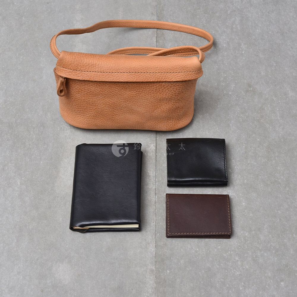 一個淺棕色的皮革斜背包，下方擺放著一個黑色皮夾、一個黑色筆記本和一個深棕色卡夾。