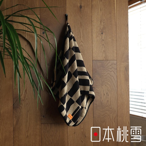 日本桃雪品牌的 SOLU 今治戶外格紋棉柔迷你浴巾，掛在木質牆面上，旁邊有綠葉植物，下方有日文文字。