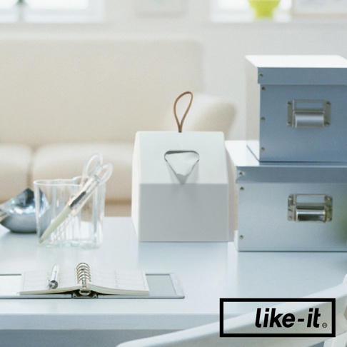 白色小屋造型衛生紙盒，頂部有皮革提把，側面有菱形開口，旁邊擺放著兩疊藍灰色收納盒。
