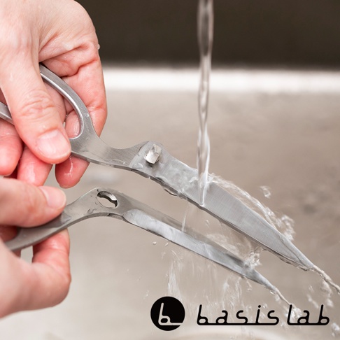 一雙可拆式不鏽鋼萬用料理剪刀，其中一把正在水龍頭下沖洗，刀刃呈現銀色金屬光澤，手部握持剪刀的動作清晰可見。