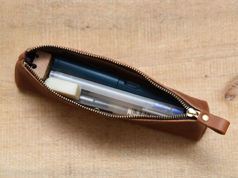 職人手作棕色皮革三角收納筆袋，拉鍊部分為金色，袋內裝有藍色筆、白色筆和橡皮擦。