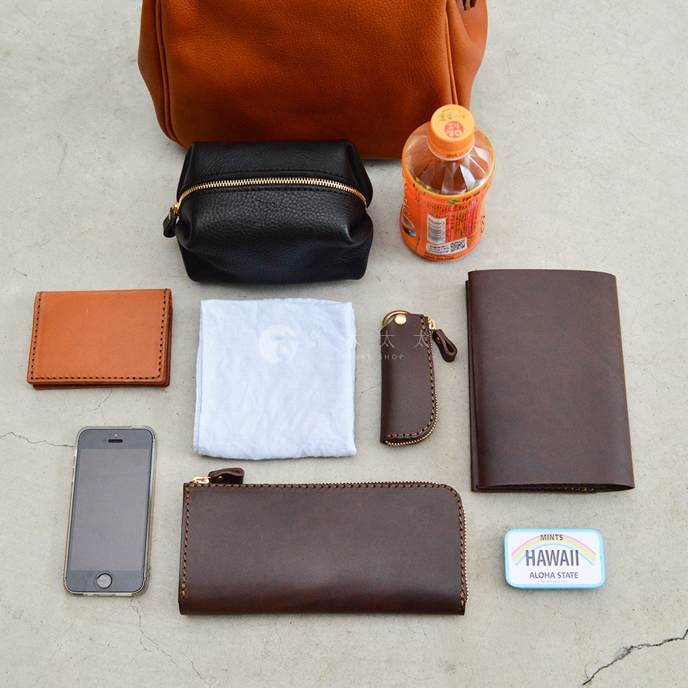 職人手作皮革Organ兩用方形口金托特包 G-55-H，展示皮革小物：棕色卡夾、黑色化妝包、淺藍色與棕色筆記本、棕色鑰匙包、長型棕色拉鍊皮夾、白色布巾、手機，以及一瓶飲料和夏威夷糖果盒。