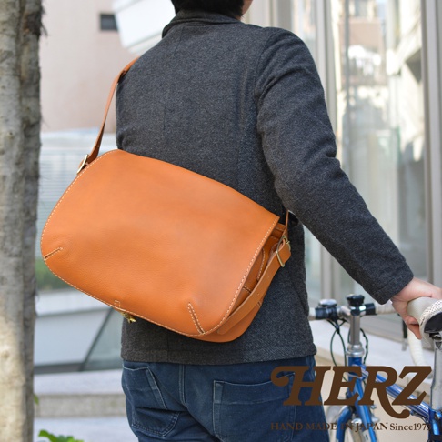 職人手作皮革復古翻蓋郵差包，採用經典棕色皮革，搭配白色縫線，側邊有金屬釦與皮帶調節。包包容量寬敞，適合日常攜帶。