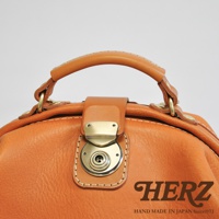 職人手作皮革後背包，採用溫暖的橘棕色皮革，搭配復古金屬旋轉鎖扣與提把，縫線精緻，展現職人細膩工藝。