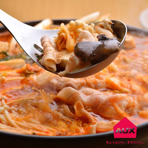 不鏽鋼濾湯火鍋杓撈起一勺豐富的火鍋料，包含豬肉片、金針菇、豆腐和香菇，湯頭呈現橘紅色，熱氣騰騰。