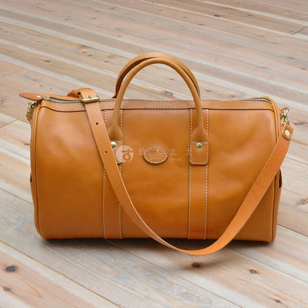 一咖棕褐色皮革製成的圓筒狀波士頓包，配有可拆卸肩背帶、提把，以及拉鍊開口。包身有白色縫線裝飾，中央有一個橢圓形皮革標誌。