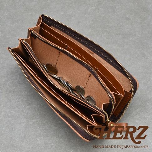 一個打開的棕色皮革長夾，內部有許多隔層，部分隔層裝有零錢。夾子的拉鍊是金色的。右下角有HERZ的標誌，下方寫著HAND MADE IN JAPAN Since1973。
