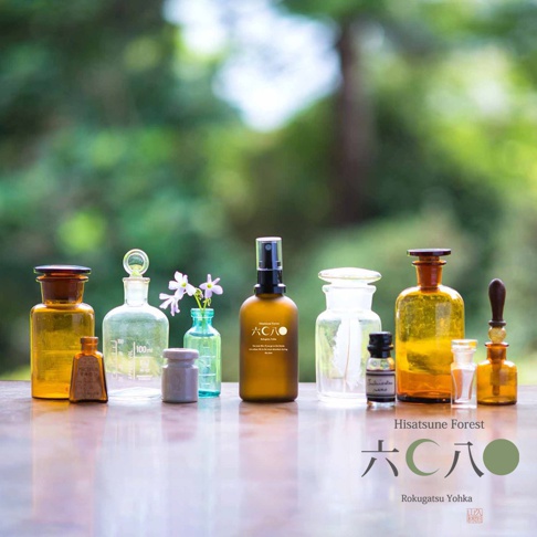 一組各式玻璃瓶裝的檜木精油產品，包含琥珀色、透明及綠色瓶身，部分瓶身帶有標籤和噴頭，其中一個瓶中插有粉紫色小花。