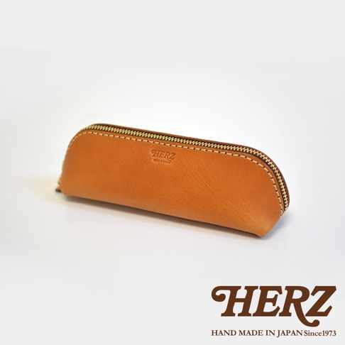 職人手作皮革三角收納筆袋，淺棕色皮革材質，搭配金屬拉鍊，筆袋表面壓印有HERZ品牌標誌。