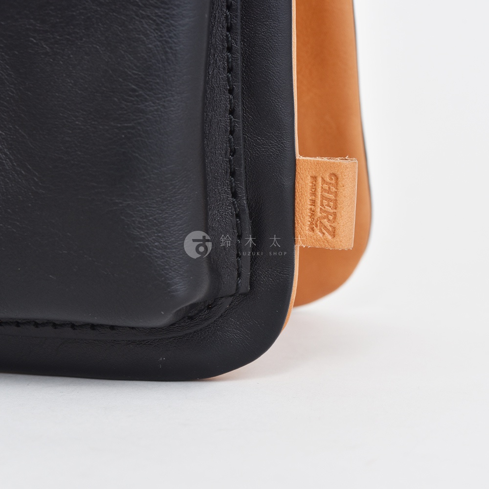 職人手作皮革翻轉雙色直式斜背包，黑色皮革搭配焦糖色皮革，右側有品牌壓印皮革標籤，標示「HERZ MADE IN JAPAN」。