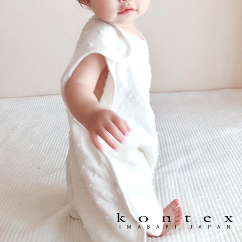 一個穿著白色無袖紗布防踢被的嬰兒，防踢被上有細小的凸點紋理，嬰兒的腳露在外面，背景是白色紋理的床鋪。