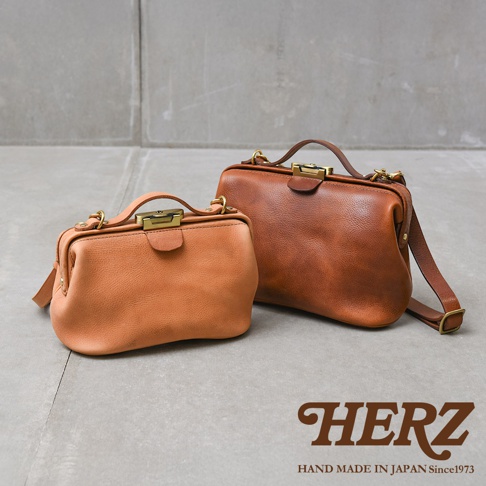 日本HERZ品牌，兩款不同尺寸的手工皮革Organ鎖孔式大開口肩背包，一淺棕色一深棕色，放置於水泥地上。