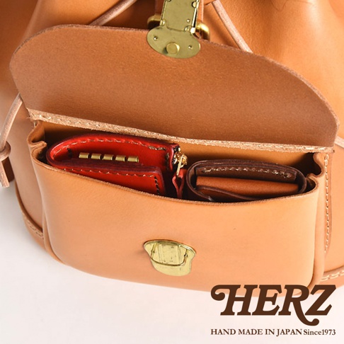 職人手作皮革後背包，特寫其主要隔層，內有紅色鑰匙包和棕色皮夾，包身為經典的淺棕色牛皮，配有黃銅色金屬扣環。