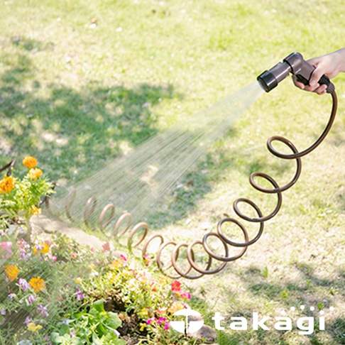 一隻手正握著一個棕色噴嘴，朝向一片綠色草地和花叢噴灑細緻的水霧。彎曲的棕色螺旋水管連接噴嘴，自然地捲曲在地面上，一端伸向草叢。