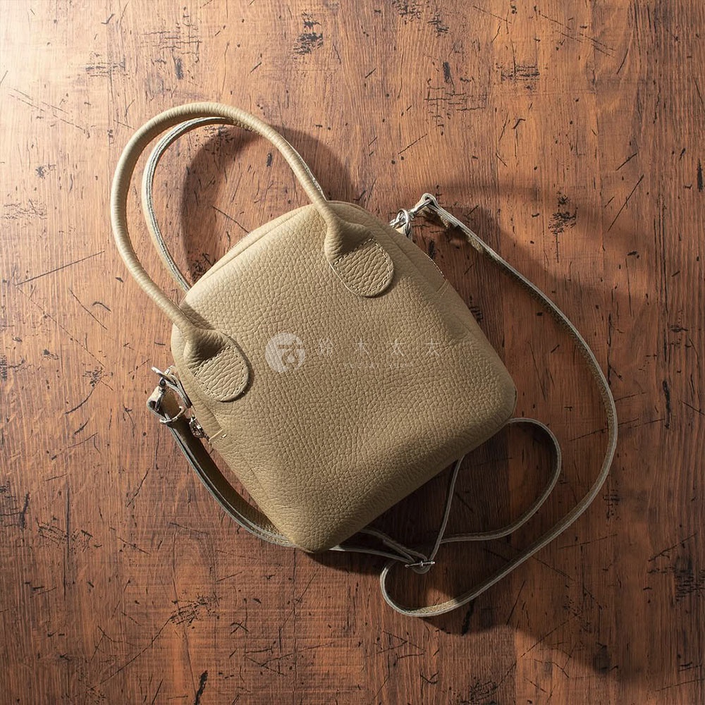 淺卡其色牛皮手提包，貝殼狀設計，配有圓形手柄和可拆卸斜挎肩帶，包身呈現細緻皮革紋理，置於木紋背景上。