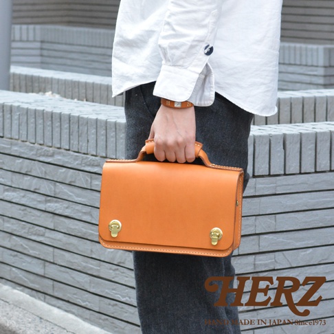 職人手作的橘色皮革迷你單肩包，帶有兩個金色雙扣和一個提把，適合手提或肩背。