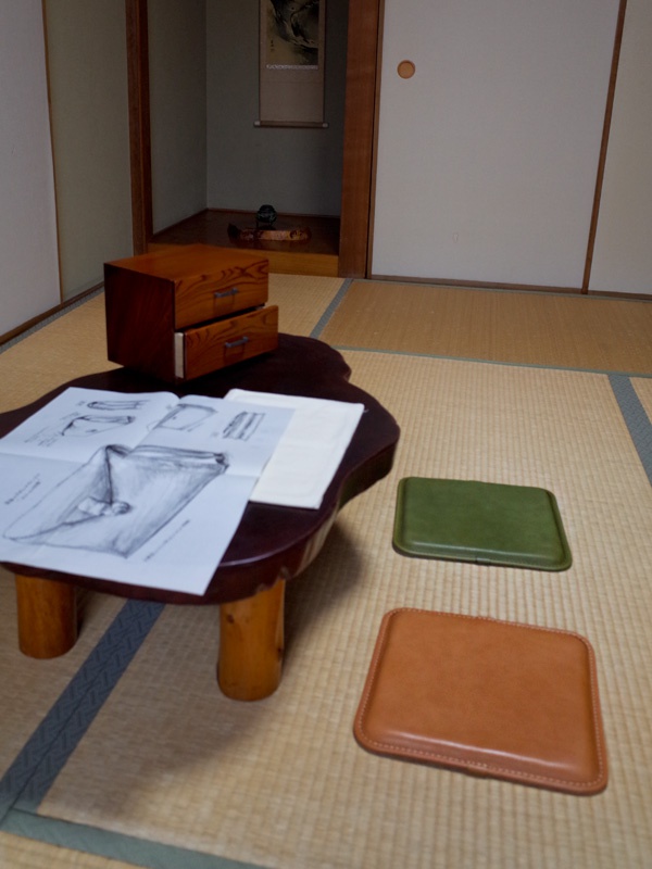 兩個職人手作皮革坐墊，一個為棕褐色，另一個為綠色，皆為方形帶有圓角的設計，邊緣有縫線。