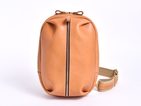 職人手作皮革立體運動斜背包，呈現自然淺棕色調。包身以光滑皮革製成，線條圓潤飽滿，中央有一條銀色拉鍊，搭配米色織帶和皮革拼接的斜背帶，整體風格簡約實用。
