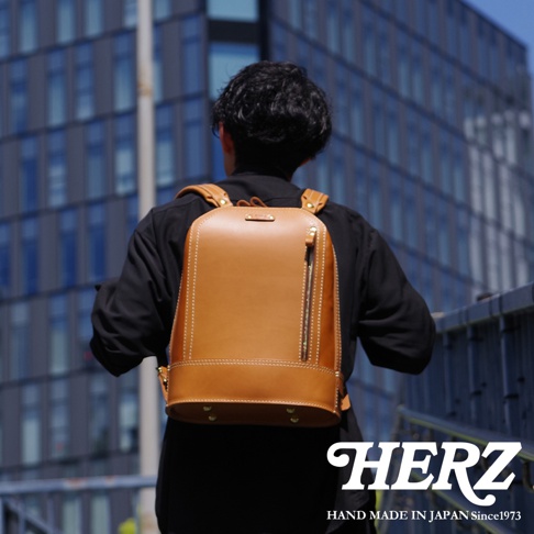 職人手作皮革立體商務後背包，採用鞣製皮革，呈現自然棕色調，雙肩帶設計，前方有拉鍊口袋，底部有金屬腳釘，整體風格簡約而具質感。