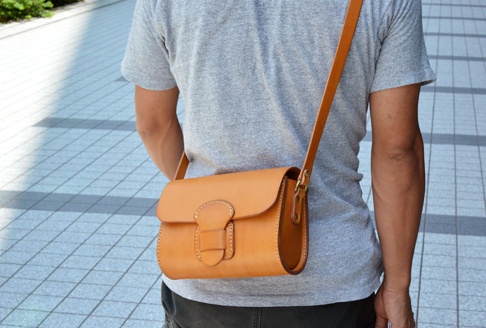一個身穿灰色T恤的人，斜肩揹著一個棕褐色皮革馬鞍包，包包上有寬版皮革扣帶和縫線細節。