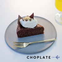 灰色的 CHOPLATE 兩用砧板盤上擺放著一塊巧克力蛋糕，蛋糕頂部覆蓋著白色鮮奶油、杏仁片和巧克力薄片，盤旁有一支金色的叉子。