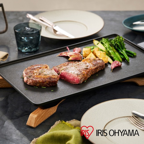 黑色輕薄電烤盤上擺放著煎好的牛排、馬鈴薯塊、蘆筍和蒜瓣，旁邊有水杯和餐具。