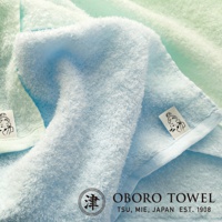 兩條觸感柔軟的純棉毛巾，一條為淺藍色，另一條為淺綠色。淺藍色毛巾疊在淺綠色毛巾之上，展示其蓬鬆的質地。兩條毛巾均帶有印有女性臉部圖案的白色標籤，以及「OBORO TOWEL」品牌標誌。