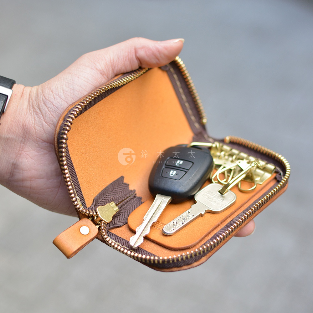 這是一張職人手作皮革橢圓形拉鍊鑰匙包，內有六鉤，圖片顯示鑰匙包裝有汽車遙控器和兩把鑰匙，包包為橘棕色，縫線為深棕色，拉鍊為金色，包包的內裡也是橘棕色。