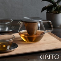 透明玻璃茶壺，內有金屬濾網，裝有琥珀色茶飲。茶壺配有銀色金屬蓋和手柄，置於淺色木托盤上，旁有一玻璃杯。