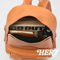 職人手作皮革休閒風後背包，展示內部空間，可見雜誌、筆記本、黑色毛衣、錢包等物品，背包為鞣製棕褐色皮革，搭配縫線與黃銅色拉鍊。