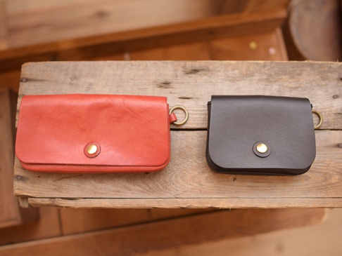 兩個職人手作皮革收納包，一個為紅棕色，另一個為深棕色，皆為長方形，帶有金色圓形按扣和金屬環，置於木質表面上。