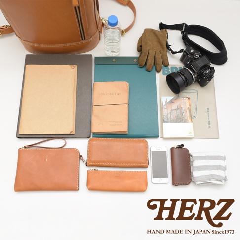 一張皮革後背包旁，擺放著棕色皮革長夾、筆狀皮夾、手機、相機、手套、筆記本、文件夾、水瓶和條紋毛巾。