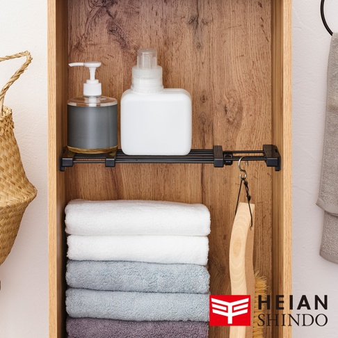 黑桿伸縮縫隙置物架，黑色金屬材質，架上放置一個透明瓶裝洗手液和一個白色瓶裝清潔劑。下方堆疊了五條不同顏色的毛巾，從上到下依序為白色、淺灰、灰藍、灰和淺紫。