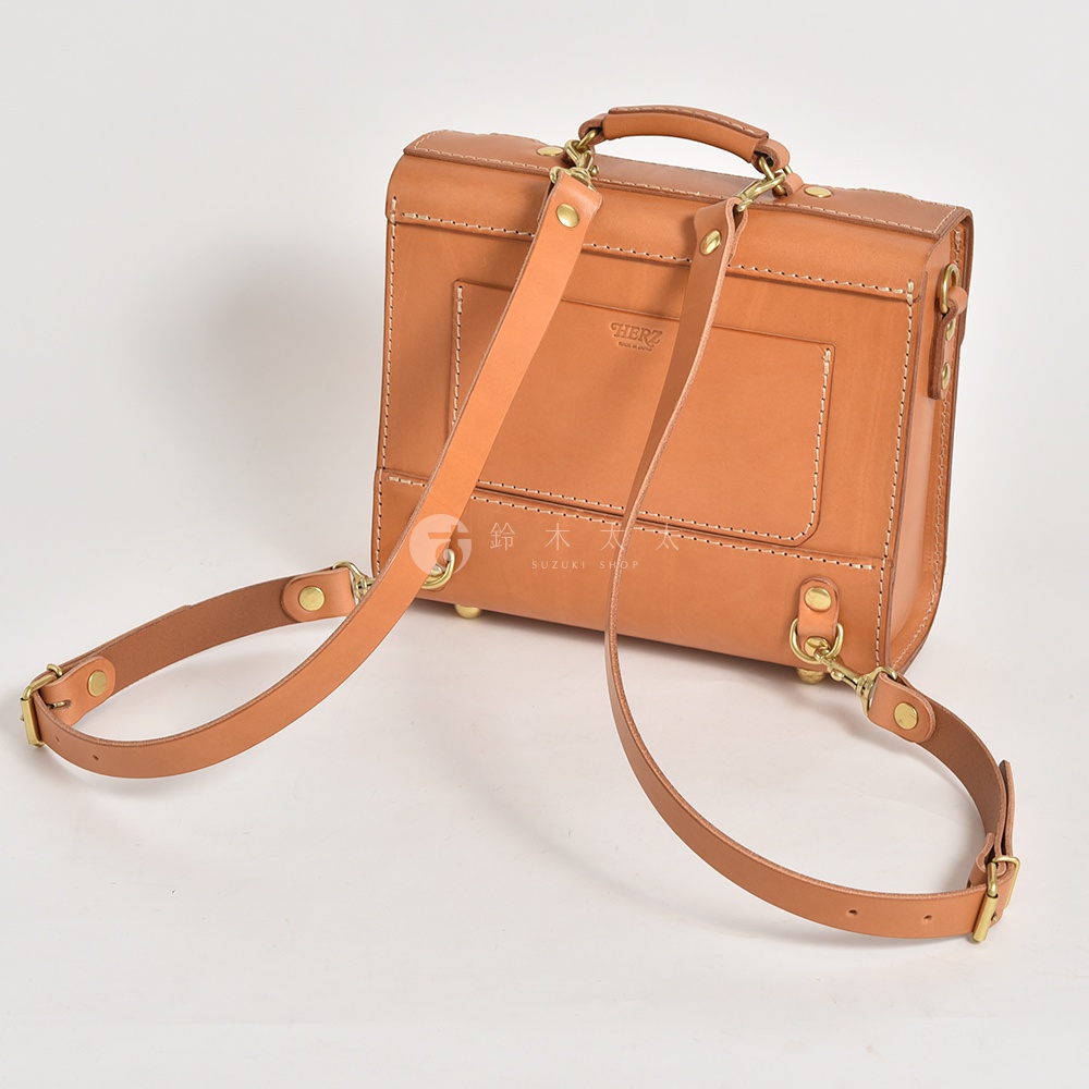 職人手作的學院風皮革三用迷你後背包，為淺棕色，材質為真皮。包款呈現方形，設有頂部提把，可見兩條可拆卸的肩帶，一條固定於包身兩側，另一條則懸掛在包身前方，配有金色金屬扣環。包身正面有兩個方形口袋，並飾有車縫線。