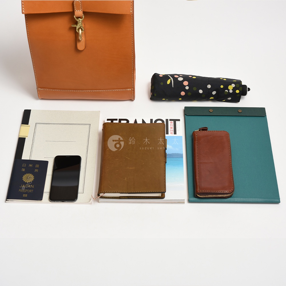 咖啡色皮革紙袋風後背包，附有金屬釦環。旁邊擺放著黑色碎花摺疊傘、日本護照、黑色手機、咖啡色筆記本、雜誌及綠色資料夾。