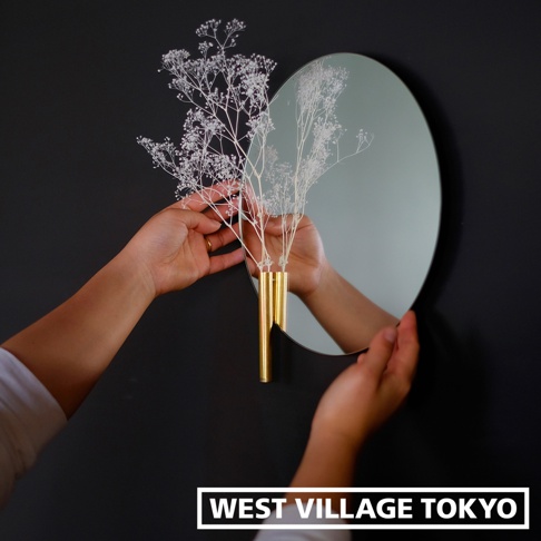 壁掛式黃銅花器，配有三個細長管狀容器，上方有白色乾燥花束，後方為圓形鏡面，鏡面反射出花器及雙手。