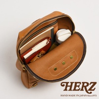 職人手作皮革大開口圓角後背包，以棕褐色皮革製成，內部裝有筆記本、皮夾和白色小物。包款設計有拉鍊開合，呈現圓潤的造型，並以白色縫線勾勒出細節。