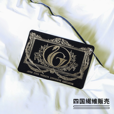一張白色光滑布料上，置放著一張黑色吊牌，吊牌上有金色浮雕的「G」字標誌，旁邊有一隻白天鵝圖案，下方印有「JAPAN DOWN PRODUCTS CORPORATIVE ASSOCIATION」字樣。