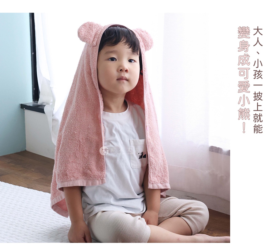 一個可愛的小男孩披著粉紅色的熊耳連帽毛巾，他穿著白色T恤和米色長褲，坐在地上。