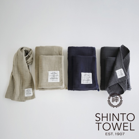 四條不同顏色的 2.5 層紗浴巾，依序為淺褐色、米白色、海軍藍及炭灰色，皆呈現織紋質地，部分浴巾疊放，並帶有白色標籤。