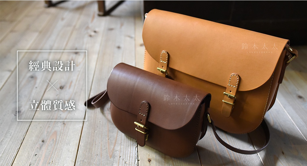 兩款職人手作皮革經典馬鞍包，一款為棕色，一款為淺棕色，皆為真皮材質，配有金屬扣環和縫線細節，置於木質地板上。
