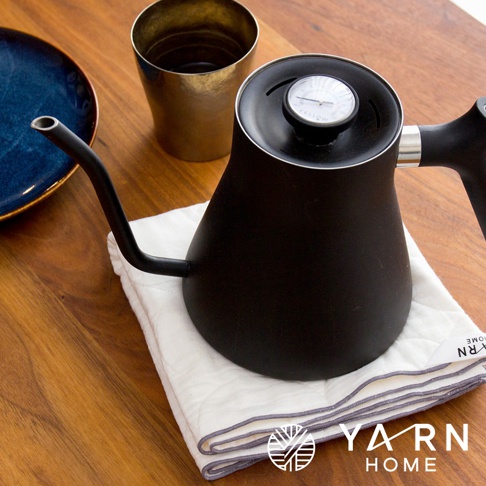 白色廚房拭巾，帶有灰色滾邊，上面放著一個黑色細嘴咖啡壺，壺蓋上裝有溫度計。