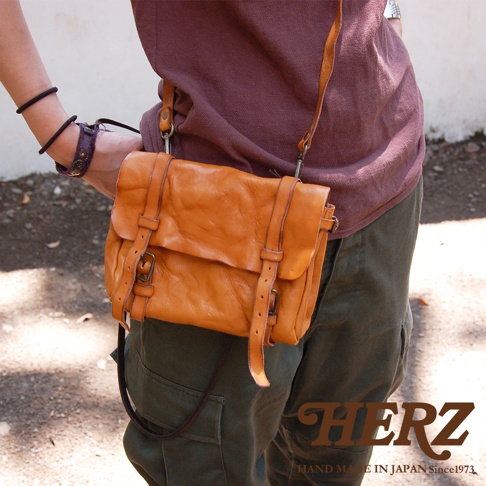 職人手作皮革水洗加工復古郵差包，呈現溫暖的橘棕色，包身為柔軟的皮革材質，搭配多條皮質束帶與金屬釦具，斜背於身。