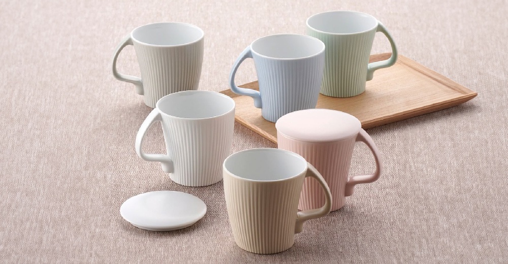 五個陶瓷馬克杯，分別為米白、淺藍、淺綠、粉紅和淺棕色，皆帶有垂直線條紋理和C形把手。其中粉紅色馬克杯配有同色系杯蓋。一個白色杯墊置於左側。