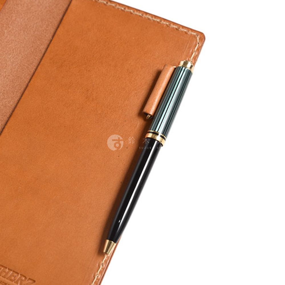 職人手作皮革輕便記事本書套，淺棕色皮革，搭配一支黑色筆身、綠色條紋筆蓋及金色筆夾的筆，筆置於書套內側的筆插上。