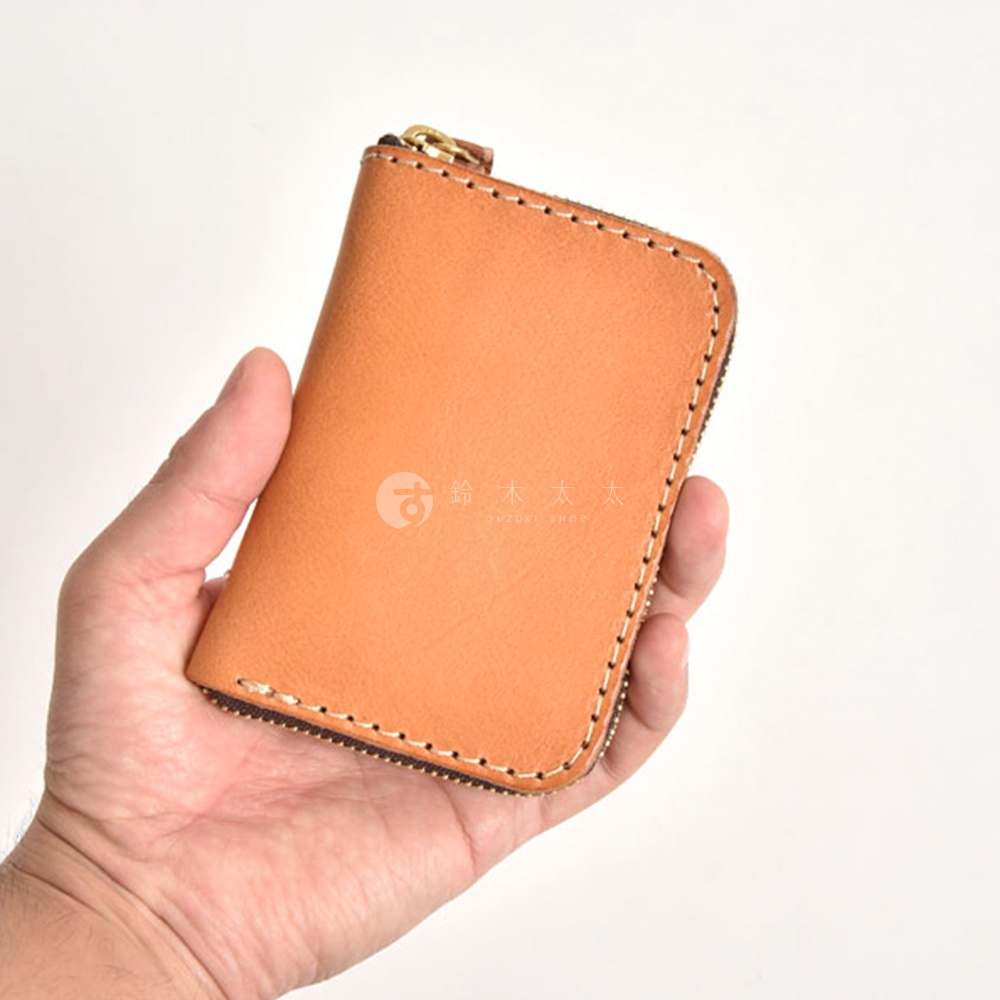 手持淺棕色職人手作皮革多用途收納鑰匙包，包體為方形圓角設計，以對比色線條縫邊，搭配金色拉鍊頭。