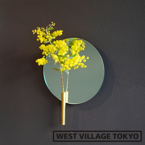 金色金屬一輪花器，瓶身直立，內插有黃色小花枝，花器固定於圓形鏡面後方，鏡面懸掛於深灰色牆面上。