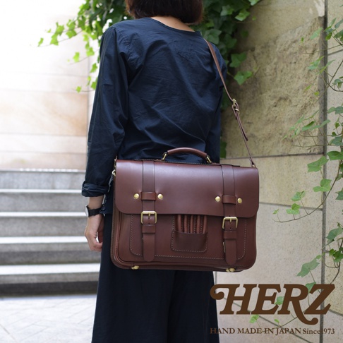 一名女子斜背著一個深棕色皮革製的歐式三用學生包，包包上有兩個皮帶扣和金屬釦，提把和背帶皆為皮革材質。