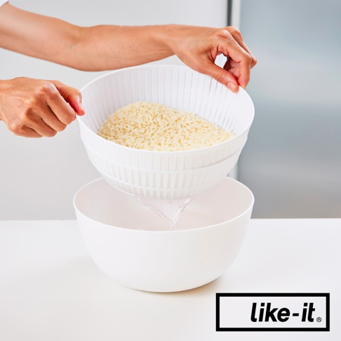 雙層白色塑膠瀝水籃，上層裝有米飯，正在被雙手提起，籃孔中水正流入下層的白色盆中。